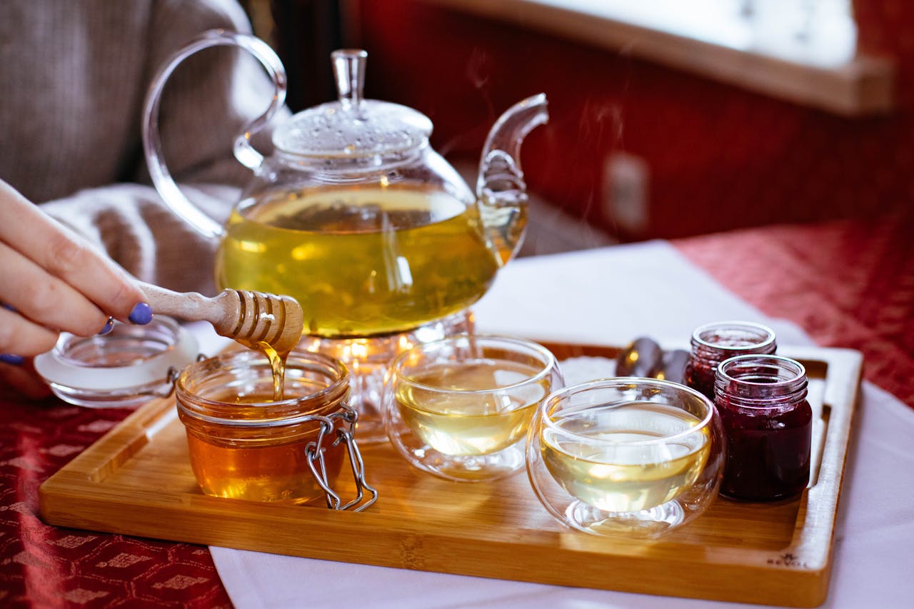 about-01 Stylish tea setup featuring honey, jam jars, and glass teapot in a warm indoor setting.